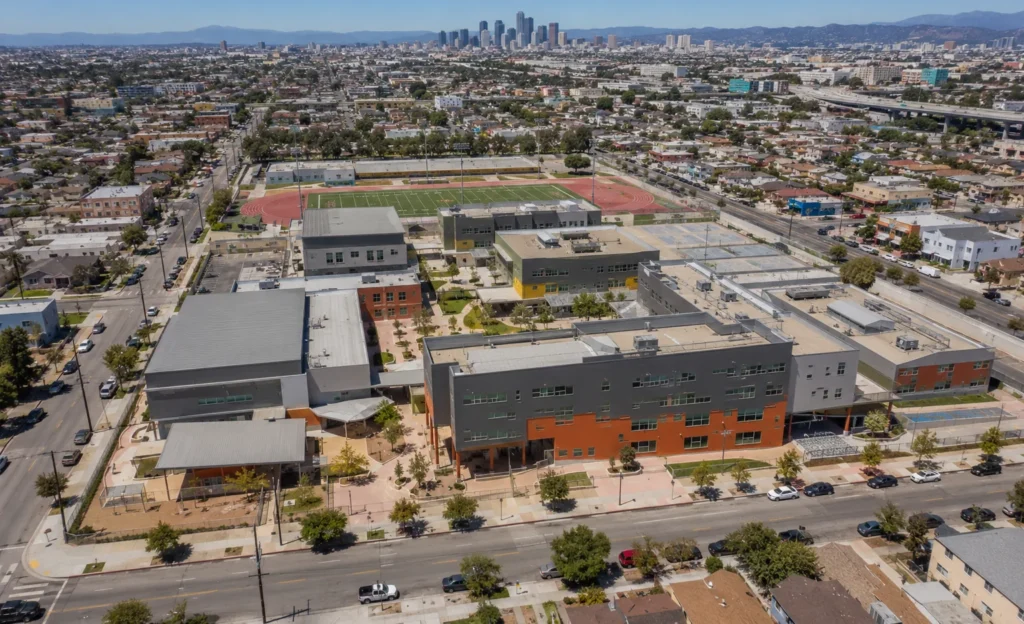 South Central Los Angeles High School No. 1 in Los Angeles, California. Aerial view of the LAUSD high school campus with classroom buildings, athletic fields, and the downtown Los Angeles skyline beyond. Los Angeles institutional education real estate.